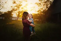 Mother cuddling her toddler in a grassy field, with golden sunset light glowing through the trees behind them.