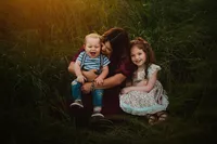 Mother cuddling her laughing toddler as her daughter sits beside them in tall grass