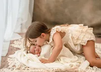 Older sister kissing her newborn sibling on a cream blanket during a natural lifestyle newborn session.