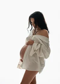 Pregnant woman looking down at her bare belly in an open white shirt, softly backlit during studio maternity portraits.