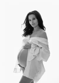 Black-and-white maternity portrait of a pregnant woman in an open white shirt, cradling her belly against a bright background.