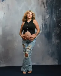 blonde mom-to-be in black vest and ripped jeans, with hands cradling a bare belly against a textured studio backdrop.