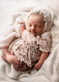 Sleeping newborn in a lace bonnet and floral outfit unposed on a cream blanket with soft lighting.