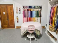 Padded newborn posing table with a pink rolling chair beneath rows of colorful photography wraps.