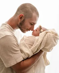 Father touching noses with his newborn, cradled in a cream blanket against soft window light.