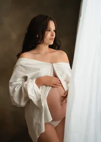 Pregnant woman in an open white shirt cradling her bare belly and gazing toward a softly lit studio window.
