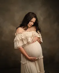 Pregnant woman cradling her belly in a flowing cream ruffled gown against a warm brown backdrop during studio maternity photos.