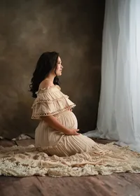 Pregnant woman kneeling in a flowing cream ruffled gown beside a softly lit window, with a warm brown studio backdrop.