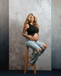 Barefoot expectant mother sitting on a wooden stool in a black top and ripped jeans during a casual maternity photoshoot.