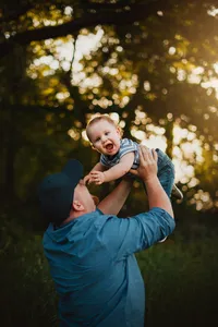 Father lifting his laughing toddler beneath leafy trees during a playful family photoshoot in golden hour light.