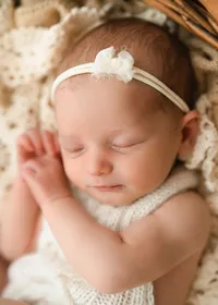 Close-up of a sleeping newborn in a cream knitted outfit and white floral headband, with her hands beside her cheek.