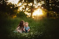 Older sister leaning close to her baby brother among tall grass and small white flowers, with sunset filtering through trees.