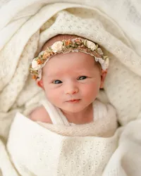 Awake newborn wearing a floral headband, swaddled in a cream knitted blanket during a natural newborn studio session.