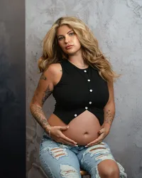 Pregnant woman seated in a black buttoned top and ripped jeans, cradling her bare belly against a textured gray backdrop.