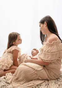 Mother holding her newborn while smiling at her older daughter beside a bright window during a lifestyle newborn photoshoot.