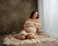 Pregnant woman seated in a cream ruffled gown, cradling her belly and gazing toward soft window light.