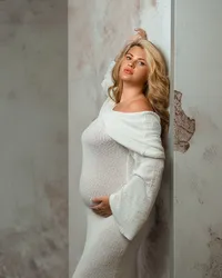 One arm raised overhead, an expectant mother wears a sheer cream gown with soft studio lighting highlighting her belly.