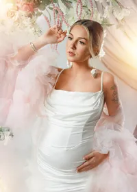 pregnant woman wearing a white satin dress and a dreamy pink flowing tulle robe beneath flowers.