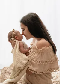 Mother touching noses with her newborn, held in a cream knitted blanket beside a softly lit studio window.