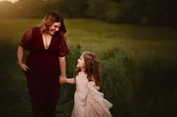 Mother and daughter smiling at each other while walking hand in hand through tall grass during family photos at sunset.