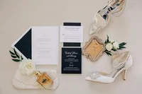 A wedding arrangement featuring invitations, a pair of white high-heeled shoes, a boutonniere, perfume, and a clutch on a neutral background.