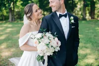 A bride and groom posing outdoors, smiling at each other, with the bride holding a bouquet of flowers.