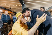 A man in a suit tenderly touches the face of a woman in a yellow outfit at a lively gathering, with onlookers in the background.