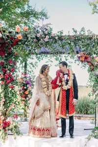 A bride and groom in traditional indian wedding attire exchange smiles under a floral archway outdoors.