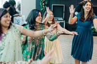 A group of women joyfully dancing together at a celebration, one in a teal dress leading with a smile.
