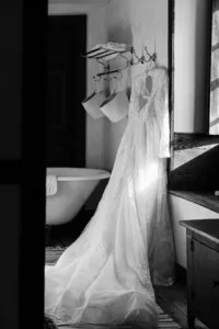 A lace wedding dress hanging beside a bathtub in a room with natural light casting shadows.