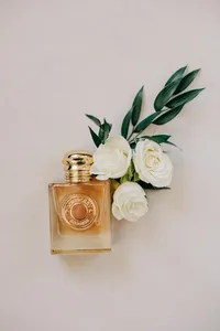 A perfume bottle with white roses and green leaves against a pale background.