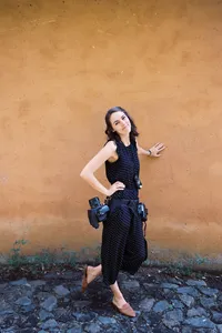 San Diego wedding photographer with two cameras hanging on her hips, leans against a textured orange wall in mexico, smiling at the camera with head tilted back wearing a black jumpsuit