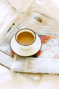 Cup of coffee resting on the morning newspaper across crisp white bedding in a guest room at L’Auberge Del Mar, hotel room detail photography