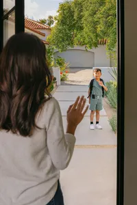 Parent waving goodbye from the doorway as her son leaves for school