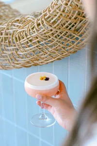 Guest holding a blush cocktail beneath a woven chair back in soft coastal light