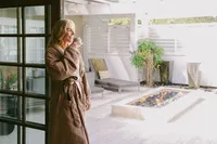 Guest in a spa robe sipping tea in the doorway of a sunlit courtyard with a fire pit and lounger after a treatment at Spa L’Auberge