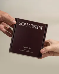 Hands holding the Soft Current Discovery Set, a deep cocoa box of four mini candles, against a warm neutral backdrop. Product packaging photography by Chelsea Loren.