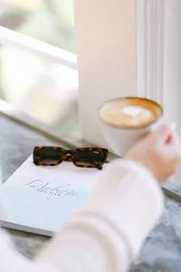 Guest holding a latte by the window beside tortoiseshell sunglasses and a branded L’Auberge Del Mar guide, coastal hotel lifestyle photography