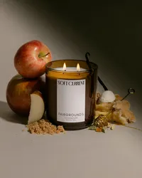 Stacked red apples beside a lit Soft Current Fairgrounds candle, with brown sugar, a honey dipper, and apple pie, lit with dramatic shadow. Editorial candle photography by Chelsea Loren.
