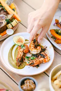 Hand reaching for grilled prawns with charred lime and herb sauce at Adelaide, surrounded by roasted vegetables, hotel restaurant food photography