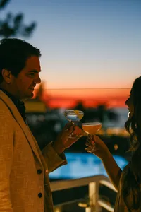 Couple toasting coupe cocktails at sunset with the glowing pool and Pacific horizon behind them at L’Auberge Del Mar, evening resort photography