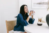 Woman holding a mug at a dining table with a cotton pad taped to her inner arm after a blood draw, diagnostics lifestyle photography