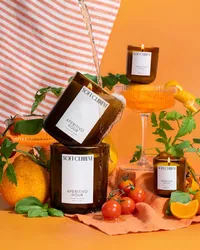 Soft Current Aperitivo Hour coconut soy candles stacked with a liquid pour splash, styled with oranges, cherry tomatoes, basil, and coupe glasses on an orange backdrop. Candle product photography by Chelsea Loren.