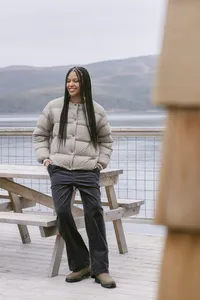 Model in a taupe down puffer and black corduroy trousers beside a picnic table on the dock