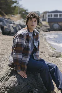Brown and blue plaid overshirt over a grey knit with denim, seated on the rocks