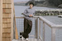 Model in a grey merino crewneck and chocolate brown wide-leg trousers on a dock railing above the bay