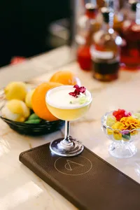 Frothy citrus sour garnished with an edible flower on a marble bar beside fresh lemons, oranges, and an Adelaide menu, hotel cocktail photography