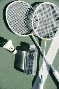 Badminton rackets, a shuttlecock, and a branded L’Auberge Del Mar water bottle casting shadows on a green court, resort amenity photography