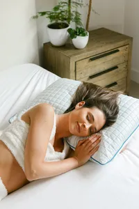 Woman asleep on a quilted cooling pillow in a styled bedroom with a wood nightstand and plants in soft morning light