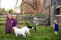 A mother in a floral dress holds a golden chicken, surrounded by her two daughters in a farmyard, with one in a purple dress and the other in blue, interacting with goats and chickens in a rustic setting during autumn in Farmington Hills